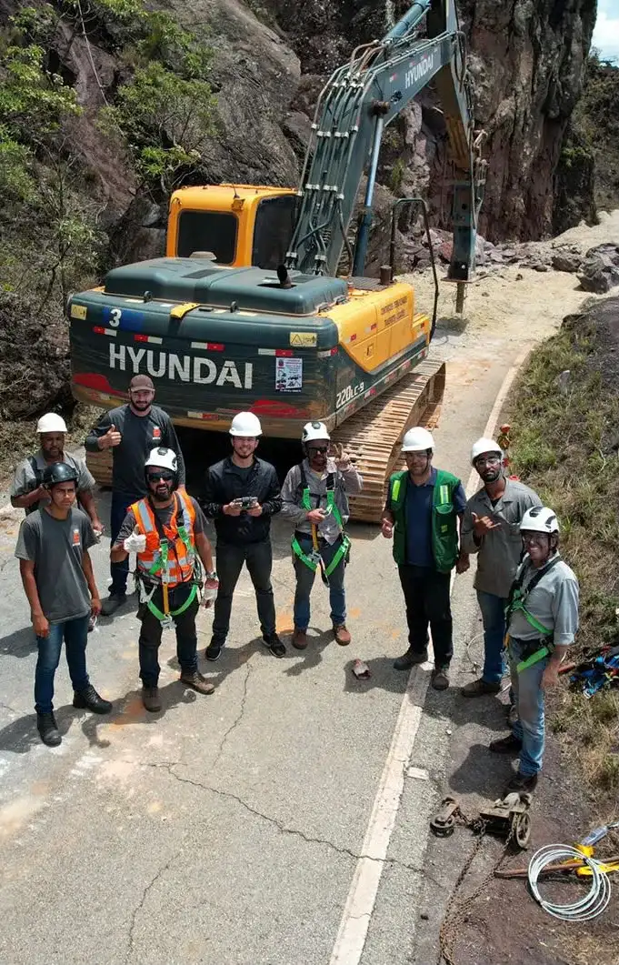 Equipe com capacete e colete ao lado de escavadeira Hyundai em estrada de serra