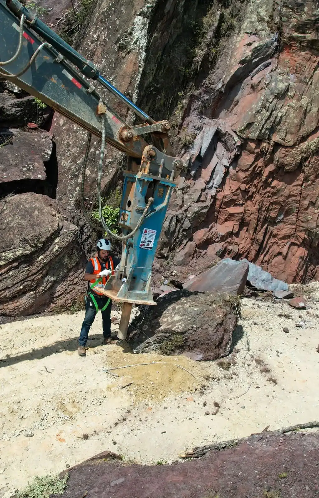 Rompedor hidráulico quebrando rocha em encosta com operador de EPI ao lado